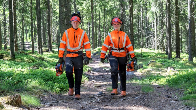Två elever med varselkläder och motorsågar går leendes på en skogsstig.