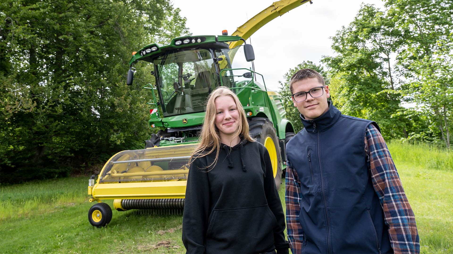 En tjej och en kille står framför en grön och gul lantbruksmaskin