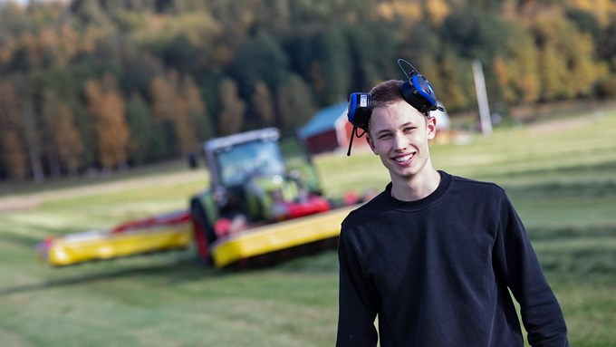 Lantbrukselev med  blå hörselkåpor på huvudet står på ett gärde med en traktor i bakgrunden.