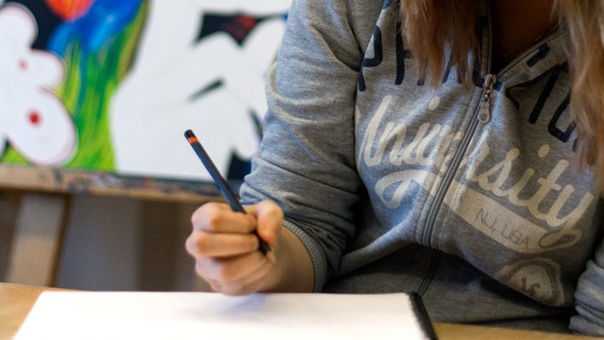 En ung kvinna håller i en penna och har ett block framför sig på bänken. Färgglad målning i bakgrunden.