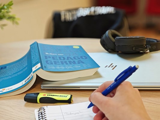 På ett bord ligger en dator, hörlurar, bok, anteckningsblock. En hand håller i en blå penna och skriver i blocket.